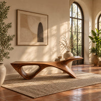 Long low wooden bench with a triangular open leg and a swept tapered end in a bright entryway with arched windows and plants