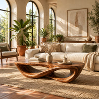 Coffee table with a twisting figure eight base and a thick oval top in a bright living room with arched windows and plants
