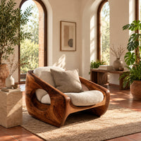 Sculpted wooden lounge chair with a circular side opening and cream cushions in a bright living room with arched windows and plants