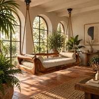 Hanging daybed swing with a sculpted wood frame and a cushioned seat in a bright porch with arched windows and plants