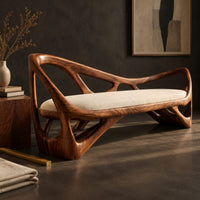 Corner detail of sculpted wooden bench with open branching sides and a padded seat beside wood, brass and linen samples