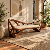 Sculpted wooden bench with open branching sides and a padded seat in a bright entryway with arched windows and plants