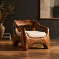 Sculpted wooden lounge chair with open arm cutouts and a cream seat cushion in a dark studio against a charcoal plaster wall