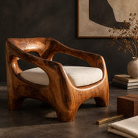 Corner detail of sculpted wooden lounge chair with open arm cutouts and a cream seat cushion beside wood, brass and linen samples