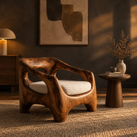 Sculpted wooden lounge chair with open arm cutouts and a cream seat cushion in a dark living room at golden hour