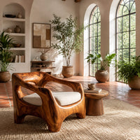 Sculpted wooden lounge chair with open arm cutouts and a cream seat cushion in a bright living room with arched windows and plants