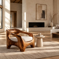 Sculpted wooden lounge chair with open arm cutouts and a cream seat cushion in a bright open plan living room