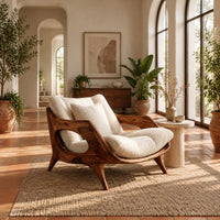 Lounge chair with a sculpted wooden frame and thick cream cushions in a bright living room with arched windows and plants