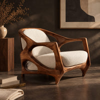 Corner detail of sculpted lounge chair with an open oval wooden frame and cream cushions beside wood, brass and linen samples