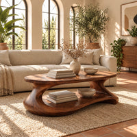 Coffee table with a sculpted pebble base and low shelf in a bright living room with arched windows and plants