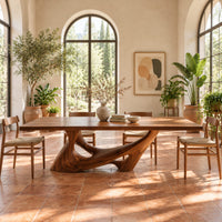 Dining table with a slab top on a sculpted pedestal with open cutouts in a bright dining room with arched windows and plants