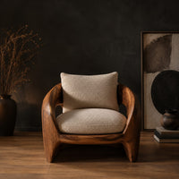 Sculpted wooden shell chair with an oval side opening and cream cushions in a dark studio against a charcoal plaster wall