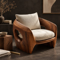 Corner detail of sculpted wooden shell chair with an oval side opening and cream cushions beside wood, brass and linen samples