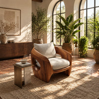 Sculpted wooden shell chair with an oval side opening and cream cushions in a bright living room with arched windows and plants