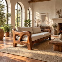 Sculpted wood sofa with open loop arms and a wavy lower rail in a bright living room with arched windows and plants