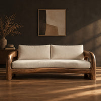 Solid wood sofa with sculpted open loop arms and a long cream seat cushion in a dark studio against a charcoal plaster wall