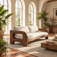 Solid wood sofa with sculpted open loop arms and a long cream seat cushion in a bright living room with arched windows and plants