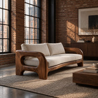 Solid wood sofa with sculpted open loop arms and a long cream seat cushion in an industrial loft living room with exposed brick