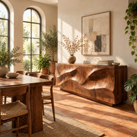 Low sideboard with wave relief across four panels and a recessed plinth base in a bright dining room with arched windows and plants