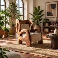 Sculpted lounge chair with looping carved arms, cutout back and cream cushions in a bright living room with arched windows and plants