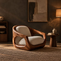 Sculpted wooden lounge chair with open sides and cream seat and back cushions in a dark living room at golden hour