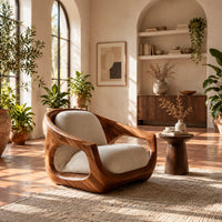 Sculpted wooden lounge chair with open sides and cream seat and back cushions in a bright living room with arched windows and plants