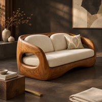 Corner detail of carved wood shell sofa with oval arm openings and a cushioned seat beside wood, brass and linen samples