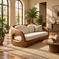 Carved wood shell sofa with oval arm openings and a cushioned seat in a bright living room with arched windows and plants