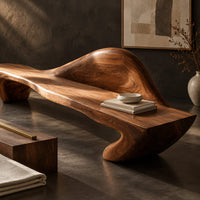 Corner detail of long wooden bench with a raised wave shaped backrest and curved legs beside wood, brass and linen samples