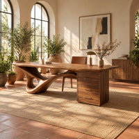 Sculptural desk with a looping wave leg and a three drawer pedestal in a bright home office with arched windows and plants