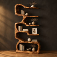 Tall serpentine bookshelf with staggered slab shelves and a loop base in a dark studio against a charcoal plaster wall