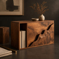 Corner detail of boxy wood side table with a carved wave front panel and an open slot beside wood, brass and linen samples