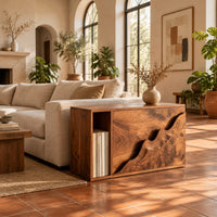 Boxy wood side table with a carved wave front panel and an open slot in a bright living room with arched windows and plants