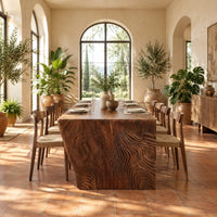 Rectangular slab dining table with tapered carved end panels in a bright dining room with arched windows and plants
