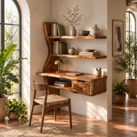 Wooden desk with a slab top, wavy side upright, two shelves and a drawer in a bright home office with arched windows and plants