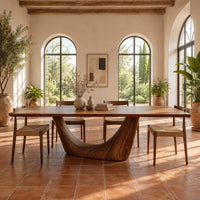Rectangular dining table with a thick top on one deep crescent pedestal in a bright dining room with arched windows and plants