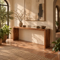 Slim waterfall console table with a smooth top and two wide slab legs in a bright entryway with arched windows and plants