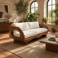 Sofa with oval arm openings, a wavy wooden frame and pale seat cushions in a bright living room with arched windows and plants