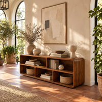 Open console table with soft edges and a long shelf above three cubbies in a bright entryway with arched windows and plants