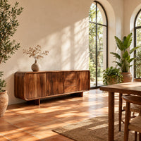 Long low sideboard with soft edges and a drawer stack between doors in a bright dining room with arched windows and plants