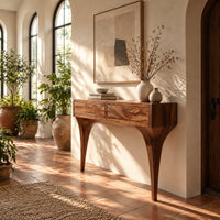 Console table with two front panels and sweeping tapered legs in a bright entryway with arched windows and plants