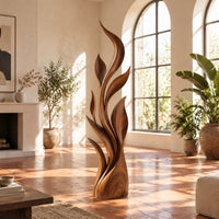 Tall flame sculpture of layered leaf blades rising from a smooth base in a bright living room with arched windows and plants