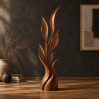 Wood sculpture of a slim leafy stem with pointed leaves and a sculpted base in a dark studio against a charcoal plaster wall