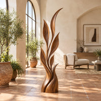 Wood sculpture of a slim leafy stem with pointed leaves and a sculpted base in a bright living room with arched windows and plants