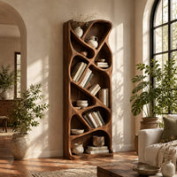 Tall narrow wooden bookshelf with slanted dividers and flowing open cubbies in a bright living room with arched windows and plants