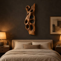 Tall carved wood wall art with oval hollows and a pointed top above a bed in a dusk lit bedroom