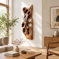 Tall carved wood wall art with oval hollows and a pointed top in a bright sunroom with green plants
