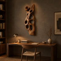 Tall carved wood wall art with oval hollows and a pointed top above a desk in a dark study corner