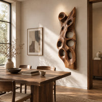Tall carved wood wall art with oval hollows and a pointed top beside a dining table in natural light