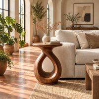 Round side table on a looped teardrop pedestal with a flat disc base in a bright living room with arched windows and plants